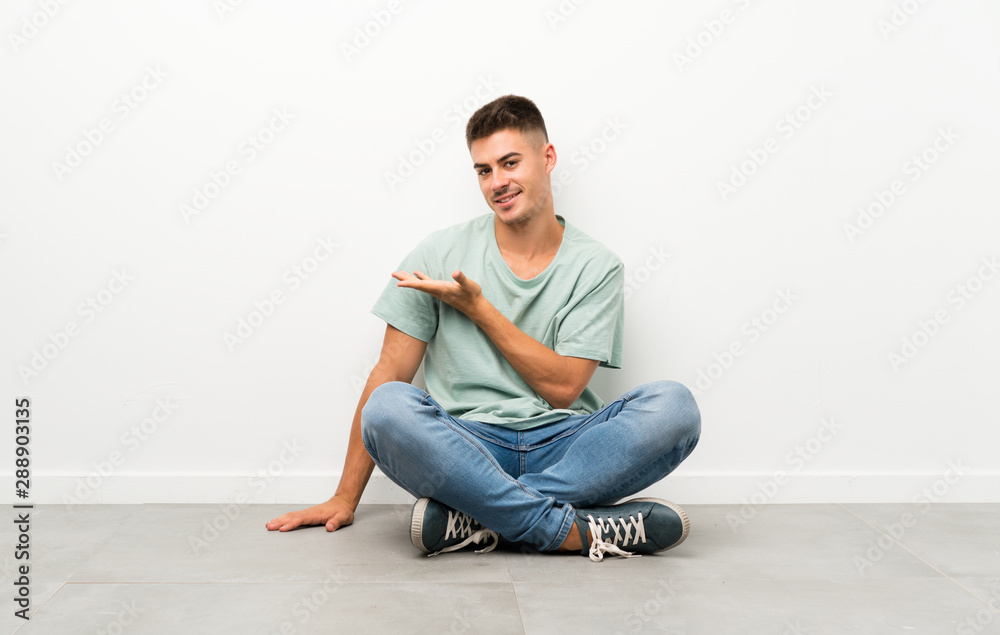 Young handsome man sitting on the floor extending hands to the side for inviting to come