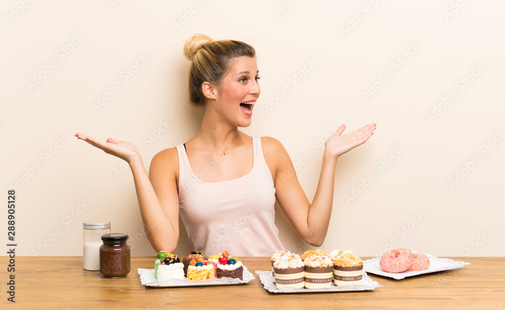 Young woman with lots of different mini cakes in a table with surprise facial expression Young woman with lots of different mini cakes in a table with surprise facial expression