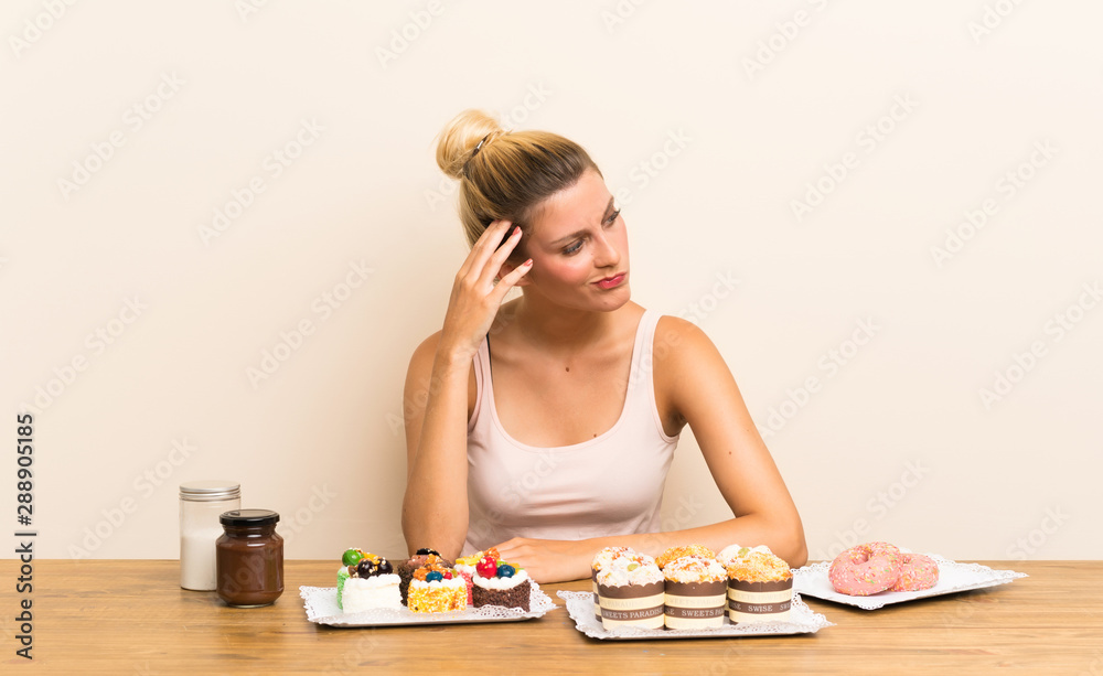 Young woman with lots of different mini cakes in a table having doubts and with confuse face expression