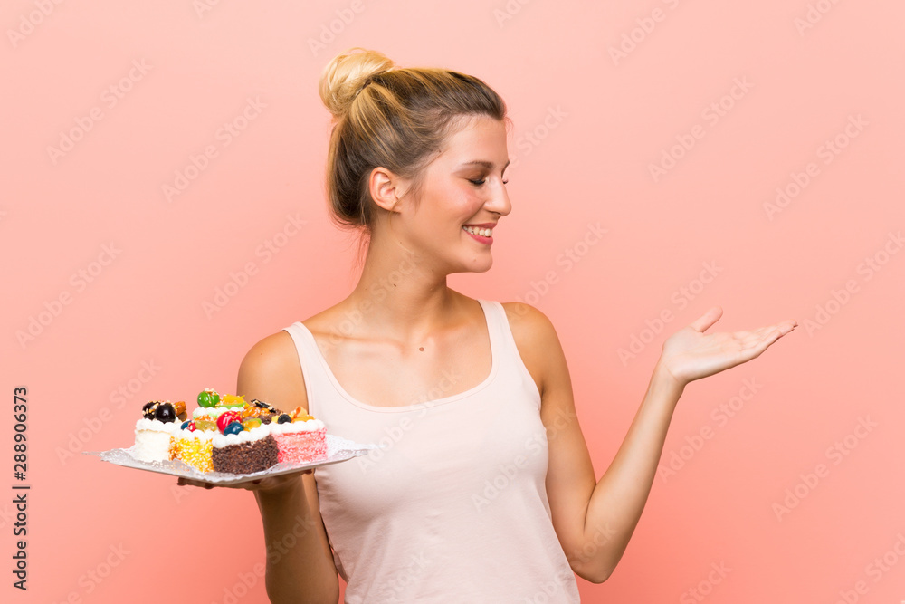 Young blonde woman holding lots of different mini cakes with surprise facial expression Young blonde woman holding lots of different mini cakes with surprise facial expression