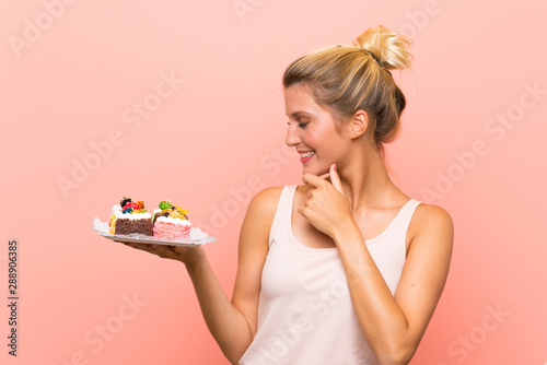 Young blonde woman holding lots of different mini cakes thinking an idea