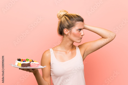 Young blonde woman holding lots of different mini cakes having doubts and with confuse face expression