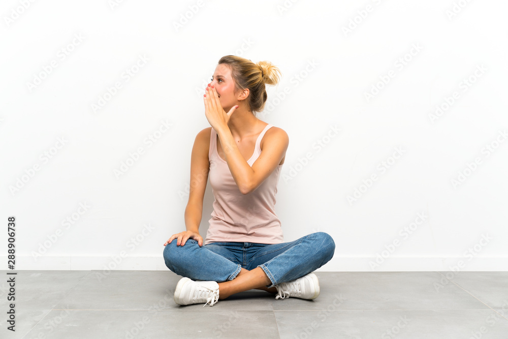 Young blonde woman sitting on the floor shouting with mouth wide open