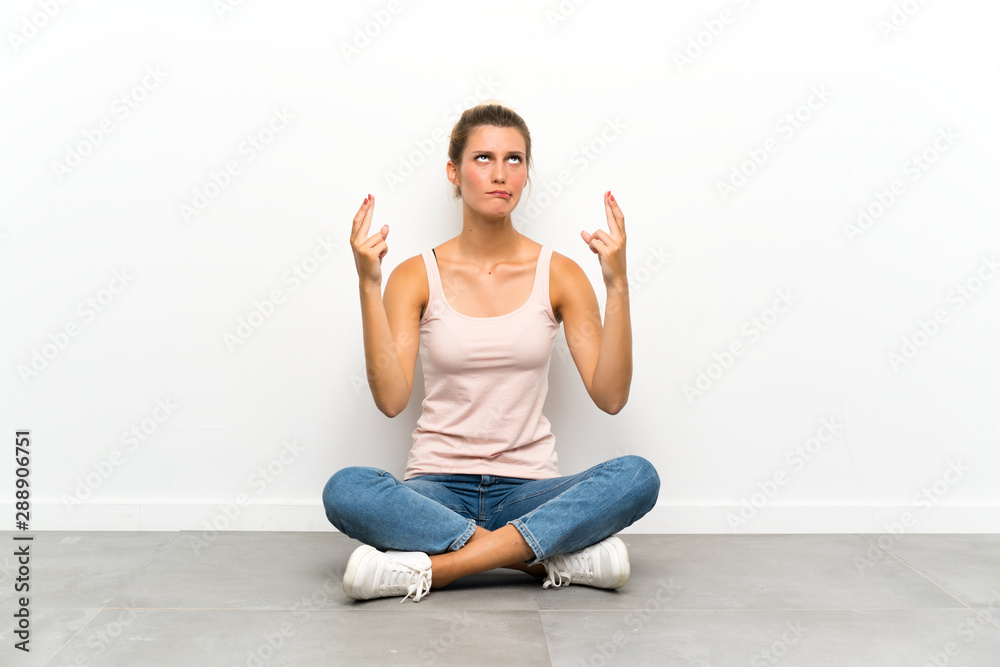 Fototapeta premium Young blonde woman sitting on the floor with fingers crossing