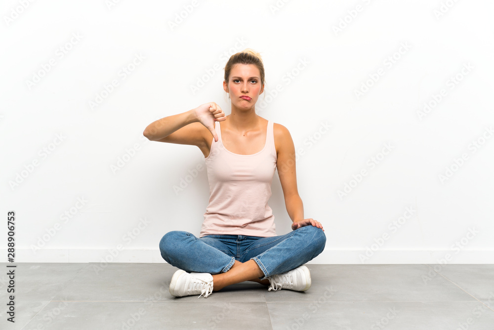 Young blonde woman sitting on the floor showing thumb down sign Young blonde woman sitting on the floor showing thumb down sign