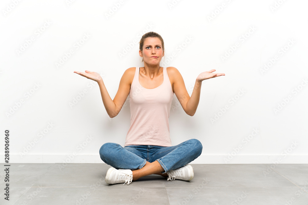 Young blonde woman sitting on the floor having doubts with confuse face expression