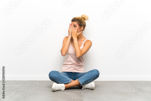 Young blonde woman sitting on the floor covering eyes and looking through fingers