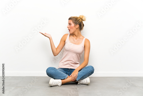 Young blonde woman sitting on the floor holding copyspace imaginary on the palm