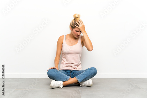 Young blonde woman sitting on the floor having doubts with confuse face expression