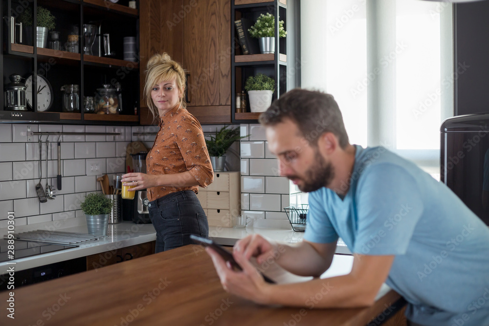 Fototapeta premium Couple in kitchen. Man is browsing on tablet while woman brings refreshments.