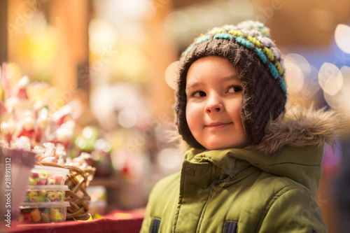 holidays, childhood and people concept - happy little boy at christmas market candy shop in winter evening