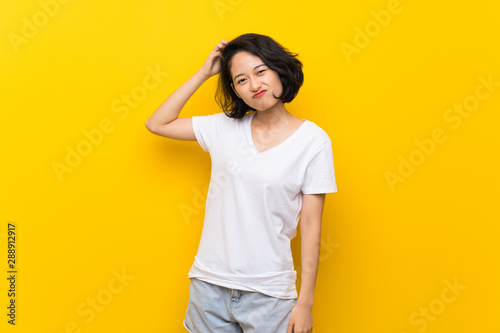 Asian young woman over isolated yellow wall having doubts while scratching head
