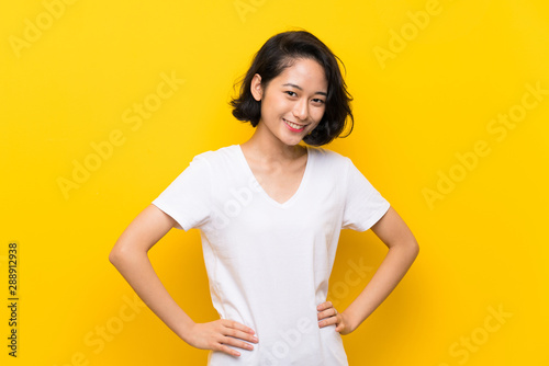 Asian young woman over isolated yellow wall posing with arms at hip and smiling