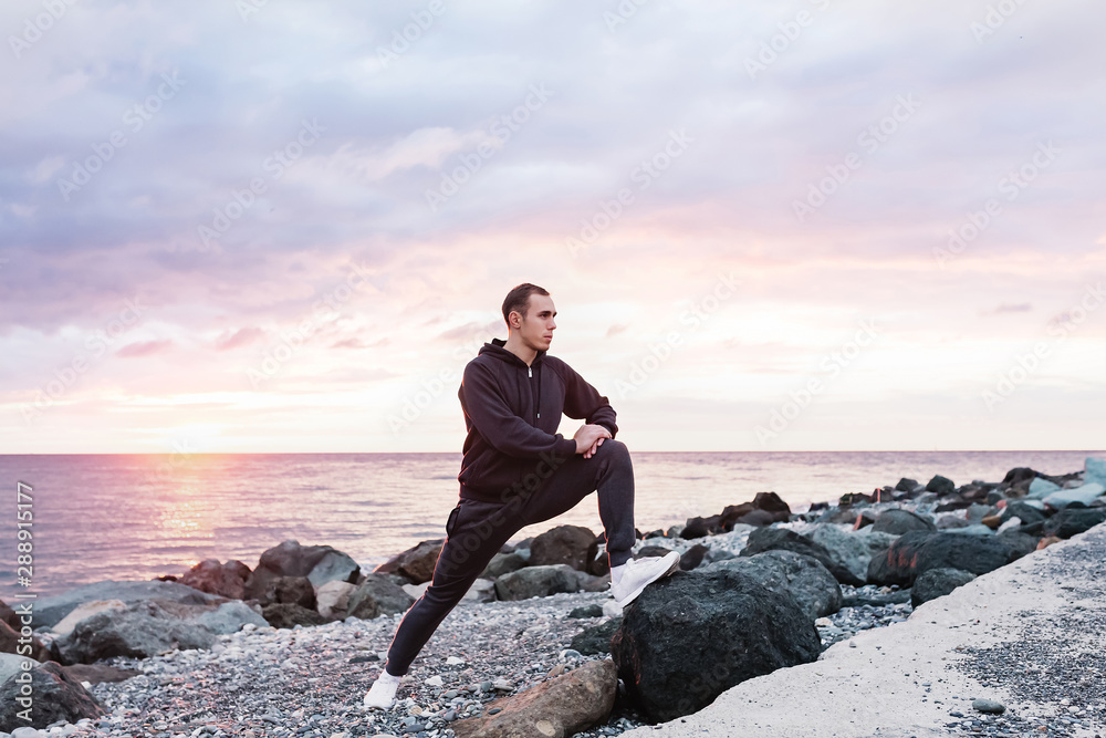 Slim tall sportsman athlete doing a warm-up on a rocky seashore against the backdrop of the sunset. Concept of sports training after a hard day. Copyspace Slim tall sportsman athlete doing a warm-up on a rocky seashore against the backdrop of the sunset. Concept of sports training after a hard day. Copyspace