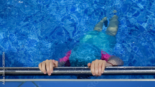Small girl in swimming cap and swimsuit dives with breath holding in pool. Child girl is diving underwater holding on the metal handrail. Unrecognizable child on swim lesson in sport school.