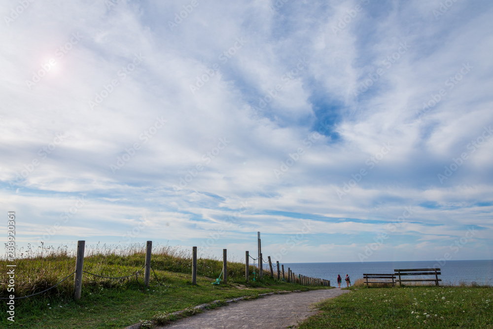 Landscape with road with two people in the background and two benches next to the coast of Comillas in Cantabria, Spain