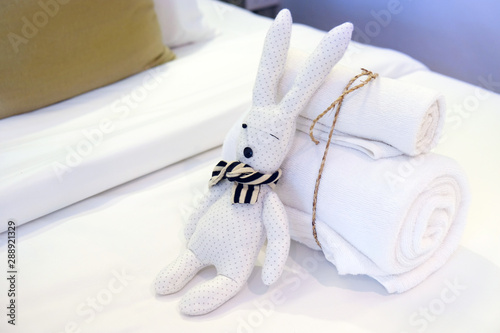 Roll of towels tied by rope, and rabbit doll on white bed sheet prepared for welcome visitor of hotel or resort