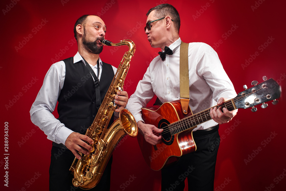 Naklejka premium Group of two musicians, male jazz band, guitarist and saxophonist in classical costumes improvise on musical instruments in a studio on red background