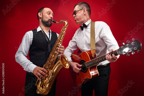 Group of two musicians, male jazz band, guitarist and saxophonist in classical costumes improvise on musical instruments in a studio on red background