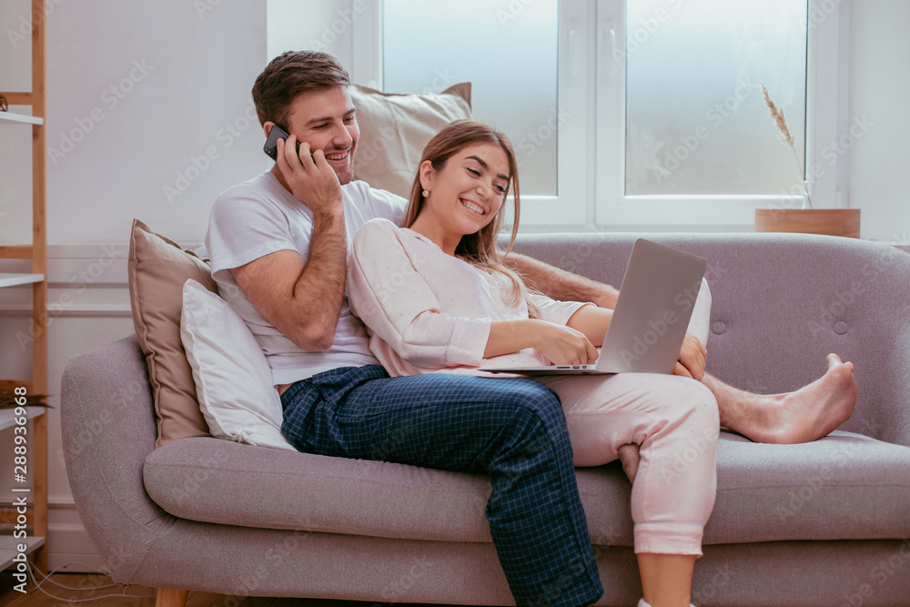 Couple in the morning making a order from the phone and watching on the notebook they are happy sitting on the sofa in pajamas Couple in the morning making a order from the phone and watching on the notebook they are happy sitting on the sofa in pajamas