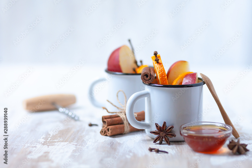 Fototapeta premium wo white metal mugs with ingredients for hot mulled wine - traditional christmas beverage. Ripe apple and orange, spices and honey. Corkscrew to open wine bottle. Holiday festive mood, rustic style