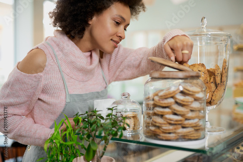 Fotografie, Obraz Attractive mixed race employee in pink turtleneck sweater and with apron opening jar with cookies
