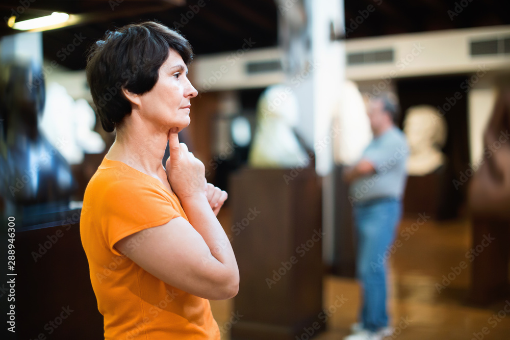 Thoughtful middle aged lady examining exposition in museum hall of ancient sculpture