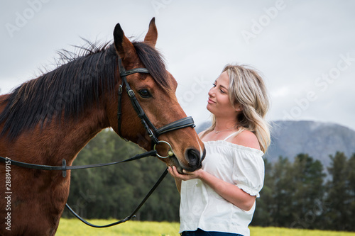 Woman standing and looking at her chestnut Arab horse interacting with him and smiling, outdoors with field of yellow flowers.