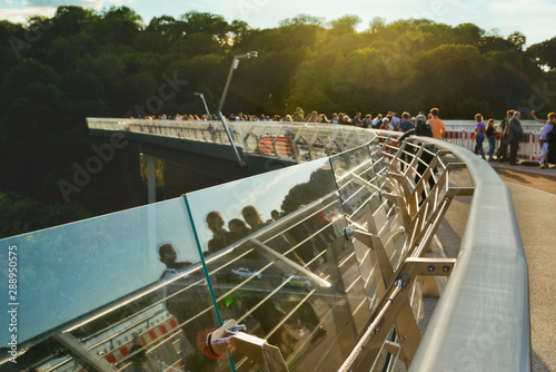 Pedestrian bridge with the transparent railing and reflected people walking through it in a summer afternoon.