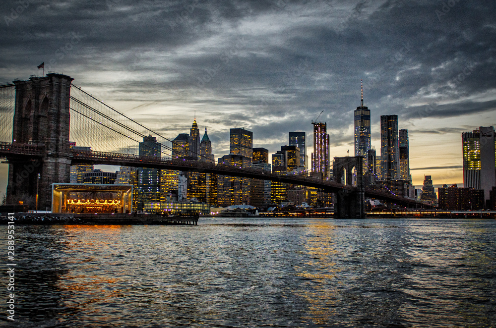 Brooklyn Bridge during a cloudy sunset in foreground with New York City skyline in background.