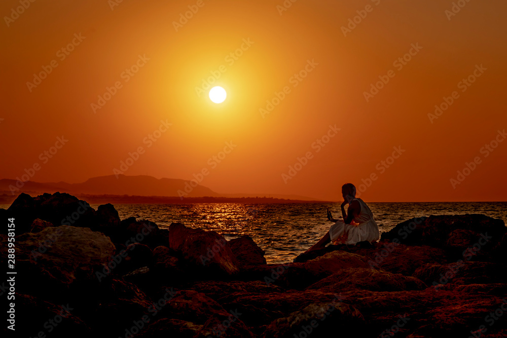 Silhouette of a young woman sitting on a stone beach and using a smartphone on a beautiful sea sunset background