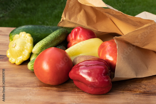 Green, yellow and red vegetables spilled out of a paper bag onto a brown wooden board against a background of green grass