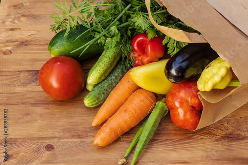 Green, yellow and red vegetables in a paper bag lying on their side on a brown wooden board, side view from above