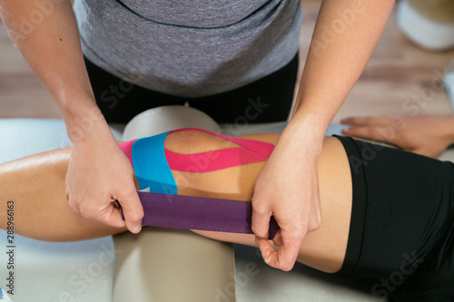 Photo detail of the hands of a physiotherapist woman gluing purple medical tape on another celestial tape and another pink one on the knee of a patient. Concept of muscle health and relaxation. photo