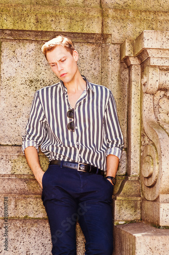Obraz na płótnie Young American Man relaxing outside in New York City, wearing blank, white striped, long sleeve shirt, blue pants, sunglasses hanging on front of shirt, standing in corner of wall, sad, thinking.. w salonie
