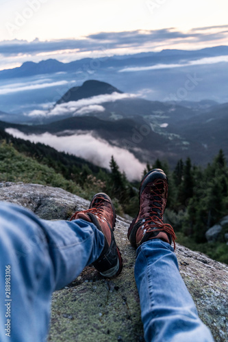 Man hiking boots enjoying wonderful breathtaking mountain view. Freedom concept. Traveller sitting and making feet selfie on beautiful landscape with forest.