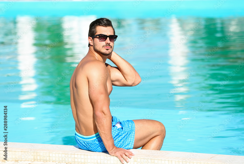 Handsome young man sitting at swimming pool edge on sunny day