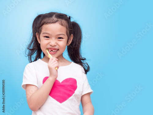 Asian little cute girl 6 years old holding a yellow toothbrush and cleans her teeth over blue backdrop.