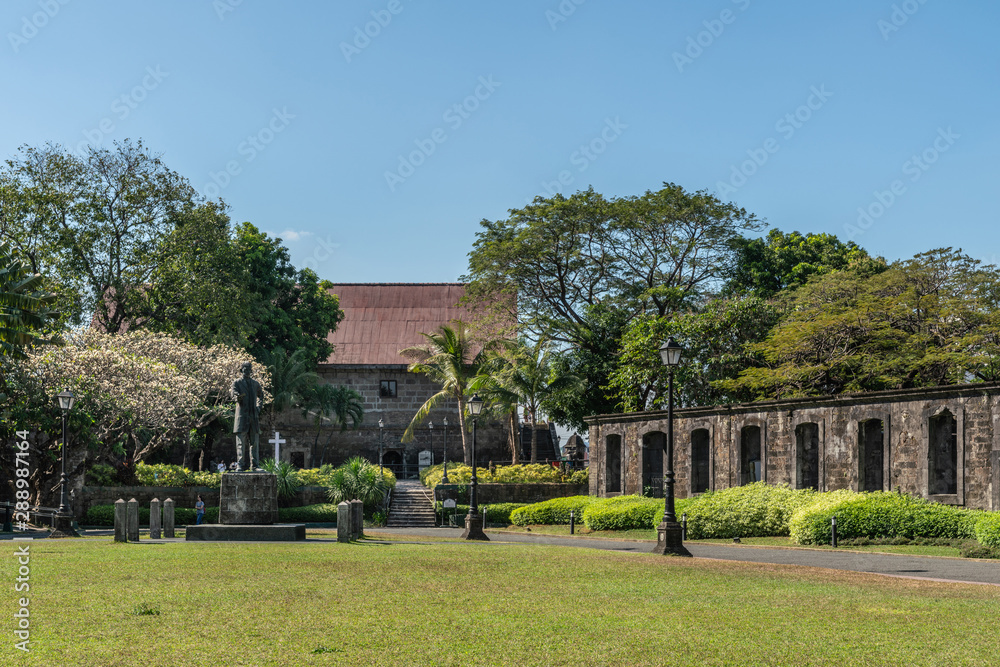 Manila, Philippines - March 5, 2019: Fort Santiaga. Green lawn of Plaza de Armas with statue of Jose Rizal. Green bushes and trees on the edges. All under blue sky.