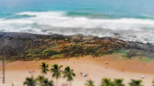 Tilt shift shot from tropical beach, with blue water, coconut trees and people walking. Villas do Atlantico, Bahia, Brazil. Drone slowly moving upwards. 