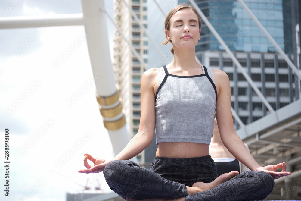 Caucasian Woman is doing yoga exercise outdoors in modern urbanistic city surrounded by tall buildings. Healthy women sitting in meditation postures with the background is skyscrapers.