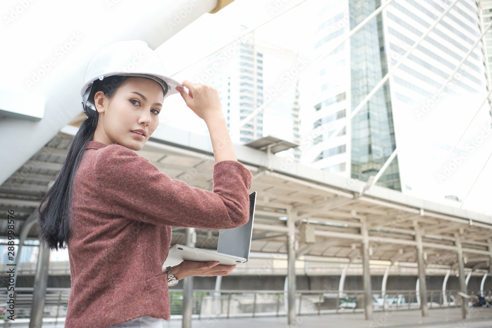 Confident Asian working woman is wearing helmet and working on laptop while standing outdoors at the walkway in Business district with high buildings. Female engineer or architect. Confident Asian working woman is wearing helmet and working on laptop while standing outdoors at the walkway in Business district with high buildings. Female engineer or architect.