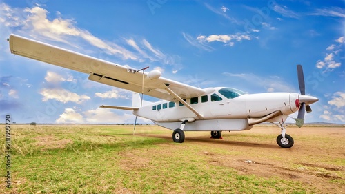 A white  single engine charter plane sits on a grass and dirt landing strip in a beautiful and remote location in Kenya
