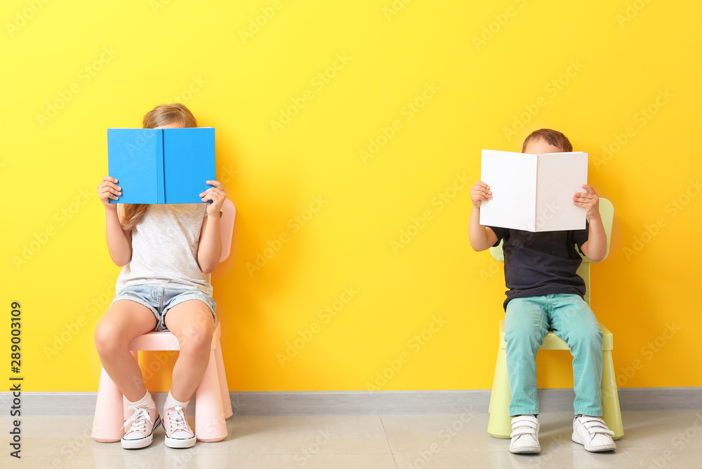 Little children with books sitting on chairs near color wall - obrazy, fototapety, plakaty 