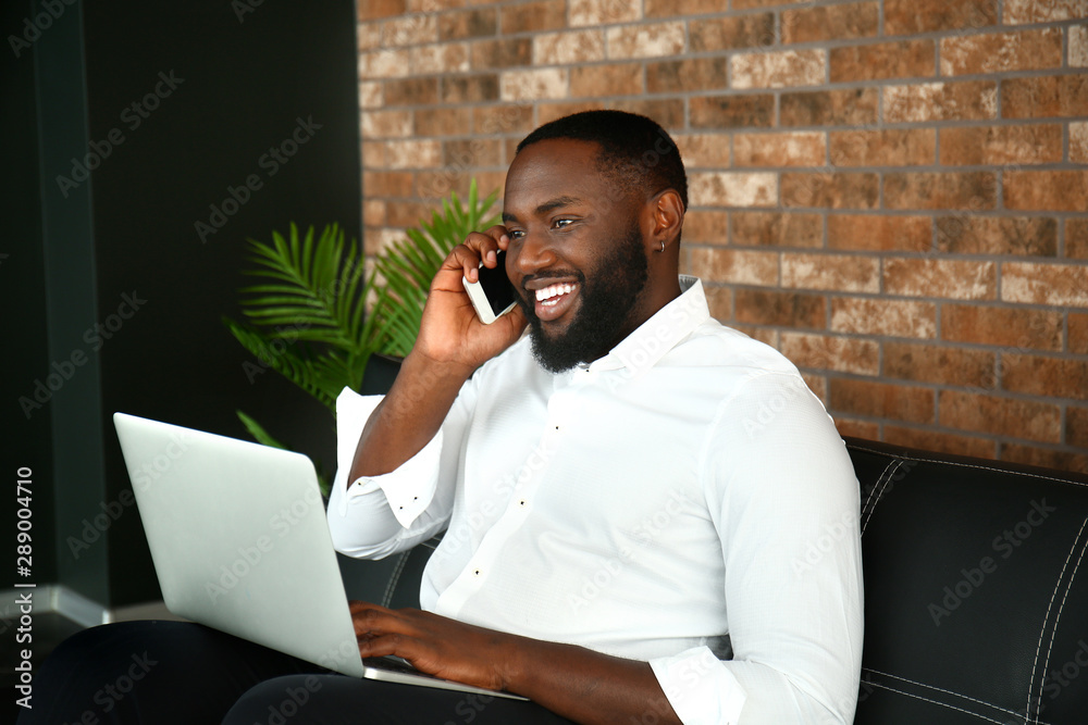 Handsome African-American businessman with laptop talking by mobile phone in office Handsome African-American businessman with laptop talking by mobile phone in office