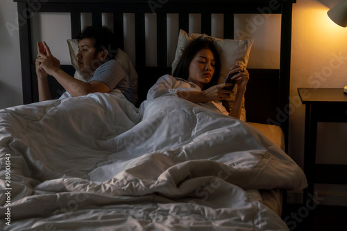 Bored young adult asian couple wife and hasband lying down on bed back to back at night don't talking with each other and using their own smartphone. Family issue and mobile phone addiction concept.