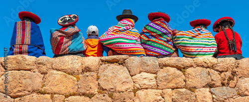 Panoramic photograph of Quechua indigenous women in traditional clothing with a boy sitting on an ancient Inca wall in Chinchero, Cusco Province, Peru.