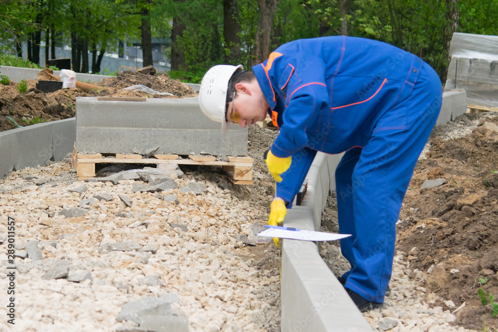 the builder, in a white helmet, blue uniform and yellow gloves, checks the progress of the laying of curb stone, the mound of rubble and leads measurements