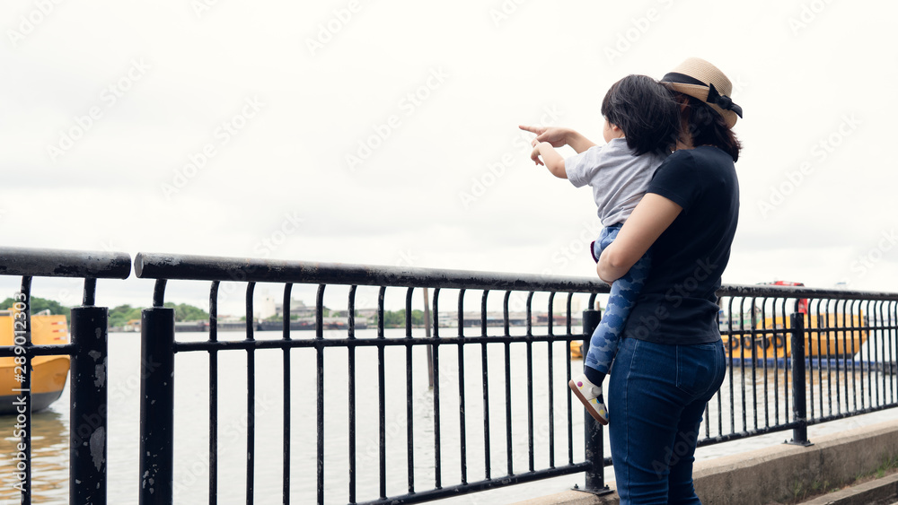 Asian mother and her toddler girl boy pointing out to the big boats in the river while answer the question from her daughter, learning and explore the environment together, happy family outing time. Asian mother and her toddler girl boy pointing out to the big boats in the river while answer the question from her daughter, learning and explore the environment together, happy family outing time.