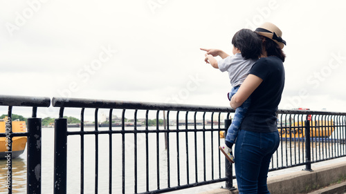 Asian mother and her toddler girl boy pointing out to the big boats in the river while answer the question from her daughter, learning and explore the environment together, happy family outing time.